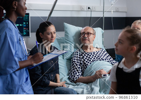 African american female physician filling out medical interventions, surgery record for senior palliative care patient. Sick elderly man family listening medication instructions from young nurse. 96861310