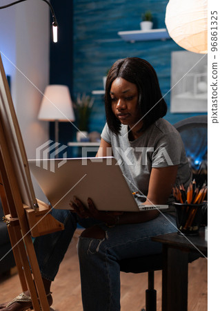 African american woman with artistic interests using laptop to take art classes via video conferencing from creative studio. Drawing student looking sketching techniques on internet to make artwork. 96861325