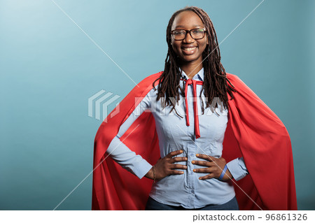 Proud and happy young superhero woman posing brave for camera while standing on blue background. Strong and selfless justice defender wearing mighty hero red cloak while standing with hands on hip. 96861326