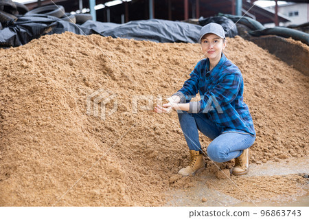 Female farmer squatting at large heap of brewer's spent grain 96863743
