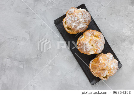 Choux Bun with whipped cream and sugar powder on top on black slate plate, gray background. Delicate choux pastry dessert. French cream puff. Top view, copy space 96864398
