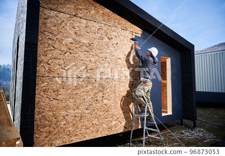 Male painter using paint roller, doing exterior paint work in a black color, standing on a ladder. Man worker building wooden frame house. Carpentry and construction concept. Male painter using paint roller, doing exterior paint work in a black color, standing on a ladder. Man worker building wooden frame house. Carpentry and construction concept. 96873053