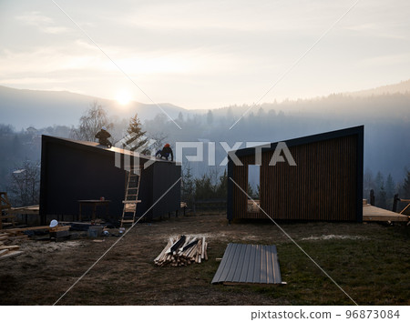 Male builders installing black corrugated iron sheet used as facade of future cottage at sunset. Men workers building wooden frame house. Carpentry and construction concept. Male builders installing black corrugated iron sheet used as facade of future cottage at sunset. Men workers building wooden frame house. Carpentry and construction concept. 96873084