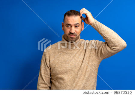 Doubtful bearded latin man thinking, scratching his head and trying to find a solution, isolated on blue background Doubtful bearded latin man thinking, scratching his head and trying to find a solution, isolated on blue background 96873530
