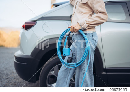 Close-up of woman holding power supply cable from her electric car, prepared for charging it in home, sustainable and economic transportation concept. 96875065