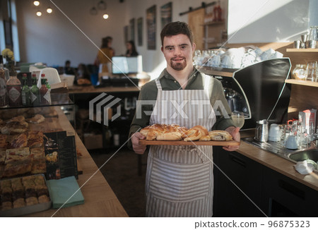 Man with down syndrom working in a cafe, carring fresh pastries. Concept of integration people with a disabilities into society. Man with down syndrom working in a cafe, carring fresh pastries. Concept of integration people with a disabilities into society. 96875323