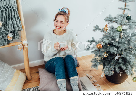 Happy Young Woman in cosy sweater drinking hot tea, relaxing on floor cusions near potted christmas tree in modern Scandi interior home. Eco-friendly cozy winter holidays. Selective focus. Happy Young Woman in cosy sweater drinking hot tea, relaxing on floor cusions near potted christmas tree in modern Scandi interior home. Eco-friendly cozy winter holidays. Selective focus. 96875925