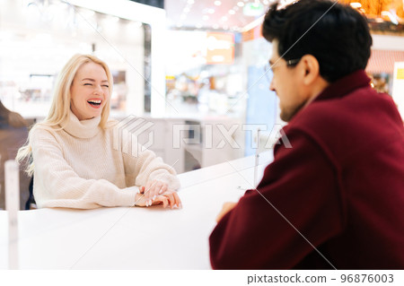 Back view from shoulder of unrecognizable young man chatting through glass partition with cheerful blonde woman standing in hall of shopping mall. Concept of lifestyle social distancing and work. Back view from shoulder of unrecognizable young man chatting through glass partition with cheerful blonde woman standing in hall of shopping mall. Concept of lifestyle social distancing and work. 96876003