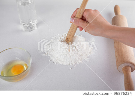 Top view of mound the flour, egg in glass, wooden spoon, water, rolling pin on white table. Dough Top view of mound the flour, egg in glass, wooden spoon, water, rolling pin on white table. Dough 96876224