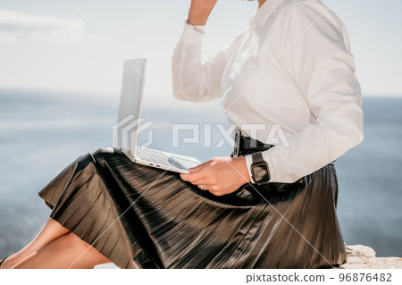 Digital nomad, Business woman working on laptop by the sea. Pretty lady typing on computer by the sea at sunset, makes a business transaction online from a distance. Freelance, remote work on vacation 96876482