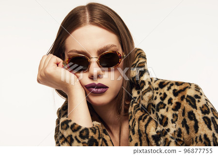 Portrait of beautiful woman posing in trendy sunglasses and animal print coat over white background. Plum colored lipstick. Bored look Portrait of beautiful woman posing in trendy sunglasses and animal print coat over white background. Plum colored lipstick. Bored look 96877755