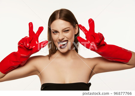 Portrait of beautiful young woman with straight brown hair and dark makeup posing in red rubber gloves over white background. Party look 96877766