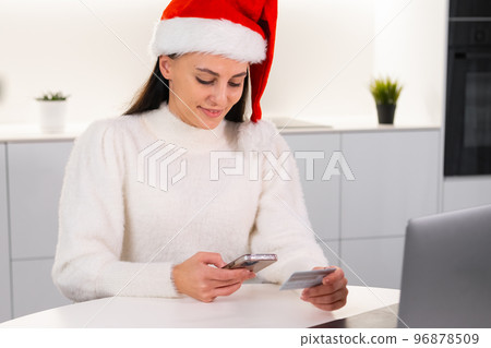 An excited woman in a red Santa Claus hat uses a credit card and mobile phone to order products for a Christmas party.  96878509