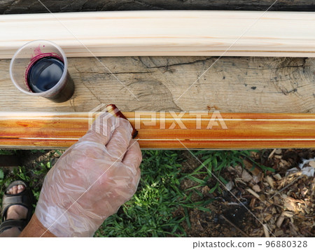 staining wooden fillets with a foam sponge and a burgundy solution of manganese in a village carpenter's workshop, a hand in a protective glove impregnates a wooden product with a sponge staining wooden fillets with a foam sponge and a burgundy solution of manganese in a village carpenter's workshop, a hand in a protective glove impregnates a wooden product with a sponge 96880328