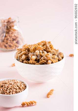 Raw pasta from buckwheat flour in a bowl and groats in a bowl on a pink background. Vertical view 96882692