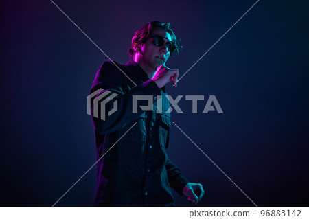 Portrait of young man in casual black shirt and trendy sunglasses, posing isolated over gradient dark purple background in neon light. Concept of human emotions, facial expression. 96883142