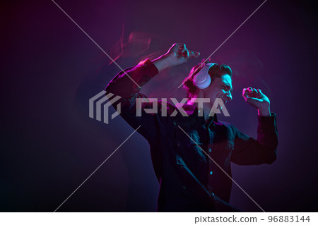 Portrait of young man in casual black shirt, dancing in headphones isolated over gradient dark purple background in neon mixed light. Concept of human emotions, facial expression. 96883144