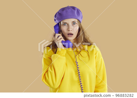 Stunned puzzled woman in purple hat and yellow sweatshirt calling phone on beige background. 96883282