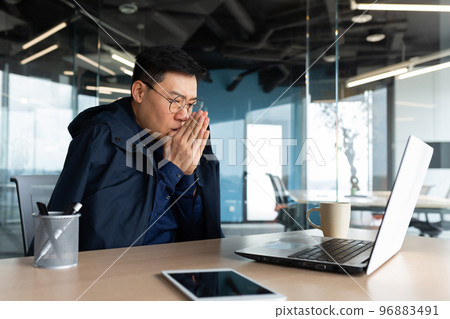 There is no heating in the office and the heater is broken, the businessman is freezing at the workplace, the man is sitting at the table inside the building using a laptop at work. 96883491
