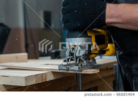 Close-up of a man cutting a wooden plank with an electric jigsaw in a workshop. 96885404