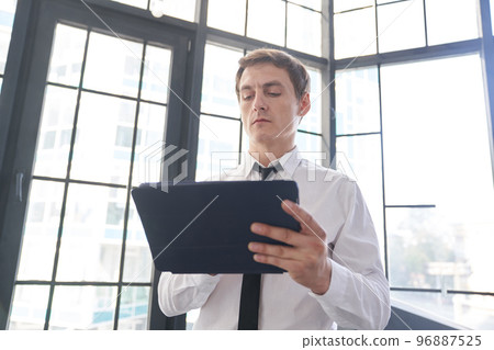 A young businessman working in an office against the background of a window. He uses a tablet with a touch screen, reading an email on it. With space to copy. High quality photo A young businessman working in an office against the background of a window. He uses a tablet with a touch screen, reading an email on it. With space to copy. High quality photo 96887525