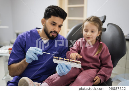Professional male dentist choosing color of filling from palette for little cute female child 96887822