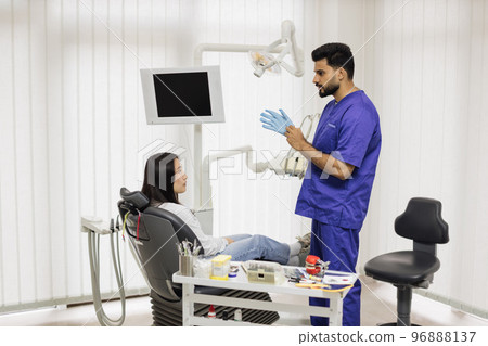 Confident bearded male dentist puts on blue rubber gloves, standing in a dental office Confident bearded male dentist puts on blue rubber gloves, standing in a dental office 96888137