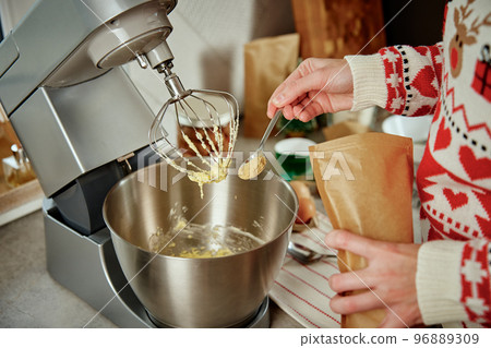 Woman in festive christmas sweater preparing dough for cookies at home kitchen. Female hands use electric mixer. Modern kitchen household appliances Woman in festive christmas sweater preparing dough for cookies at home kitchen. Female hands use electric mixer. Modern kitchen household appliances 96889309