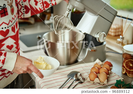 Woman in festive christmas sweater preparing dough for cookies at home kitchen. Female hands use electric mixer. Modern kitchen household appliances Woman in festive christmas sweater preparing dough for cookies at home kitchen. Female hands use electric mixer. Modern kitchen household appliances 96889312