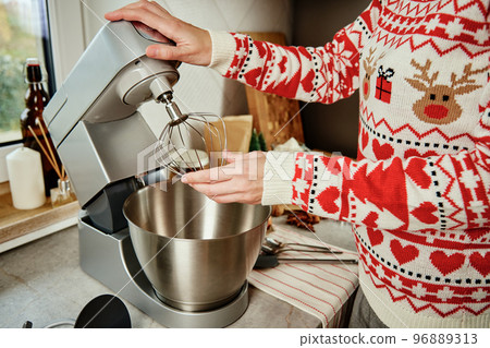 Woman in festive christmas sweater preparing dough for cookies at home kitchen. Female hands use electric mixer. Modern kitchen household appliances Woman in festive christmas sweater preparing dough for cookies at home kitchen. Female hands use electric mixer. Modern kitchen household appliances 96889313