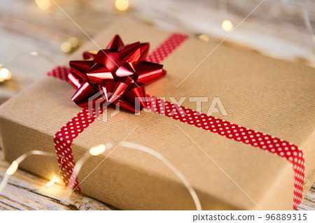 Above view of festive gift box wrapping in craft paper with festive ribbons and bow. Present for Christmas holidays on wooden background with bokeh lights Above view of festive gift box wrapping in craft paper with festive ribbons and bow. Present for Christmas holidays on wooden background with bokeh lights 96889315