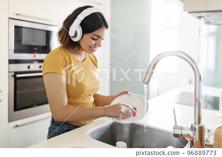 Young Arab Woman Wearing Wireless Headphones Washing Dishes In Kitchen Young Arab Woman Wearing Wireless Headphones Washing Dishes In Kitchen 96889773