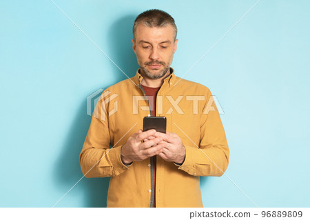 Mature caucasian man using cellphone, texting via smartphone, communicating online standing over blue background 96889809