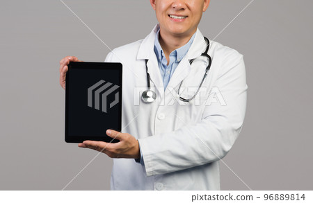 Smiling adult japanese guy therapist in white coat with stethoscope shows tablet with empty screen 96889814