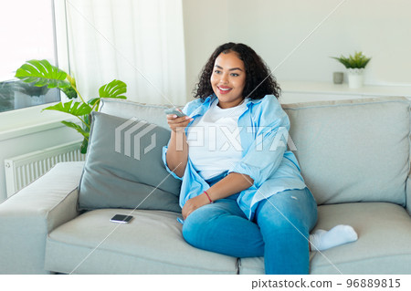 Happy african american plus size woman watching TV, switching channels with remote controller, sitting on sofa at home 96889815