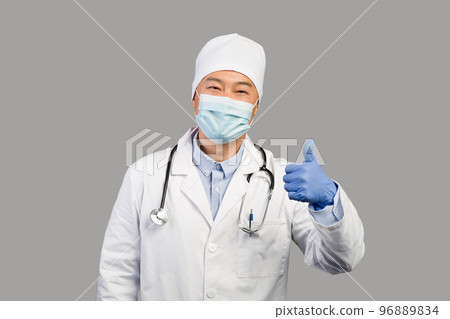 Smiling middle aged chinese male therapist in white coat, protective mask with stethoscope showing thumb up 96889834
