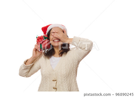 Overjoyed excited charming woman in Santa hat, covers her eyes, cutely smiles while holding a gift box with happy Christmas present, isolated on white background with copy space. Boxing Day. New Year 96890245