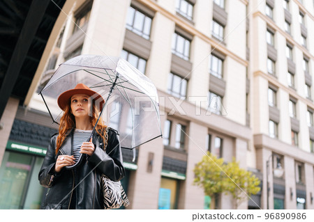 Low-angle view of charming redhead young woman in fashion hat waiting in city street holding transparent umbrella in rainy weather rain, looking away. Concept of female lifestyle at autumn season. Low-angle view of charming redhead young woman in fashion hat waiting in city street holding transparent umbrella in rainy weather rain, looking away. Concept of female lifestyle at autumn season. 96890986