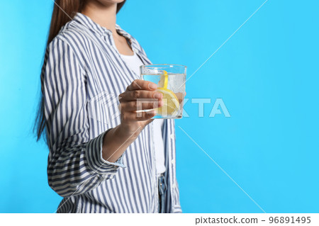 Young woman with glass of lemon water on light blue background, closeup 96891495