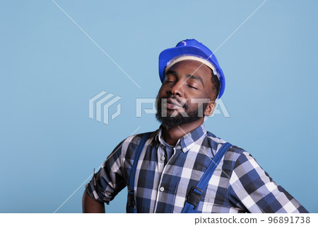 Construction worker with a dream to start work very early in the morning. African american contractor wearing hard hat and work uniform with eyes closed. against blue background, studio shot. 96891738