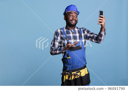 African american contractor talking on cell phone video call against blue background in studio shot. Cheerful construction worker in coveralls talking on smart phone about new work contract. 96891790