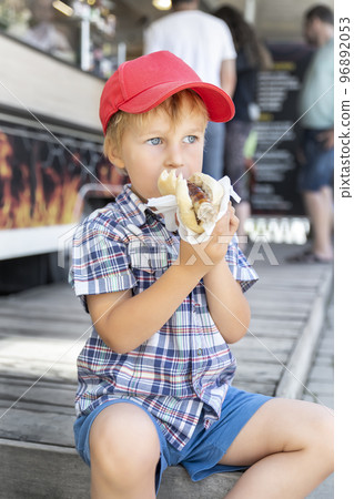 Cute adorable pensive little boy kid enjoy eating hot dog sausage in bread near street cafe stall outdoors. Child healthy eating lunch hotdog. Junk food and fast food unhealthy snack concept 96892053