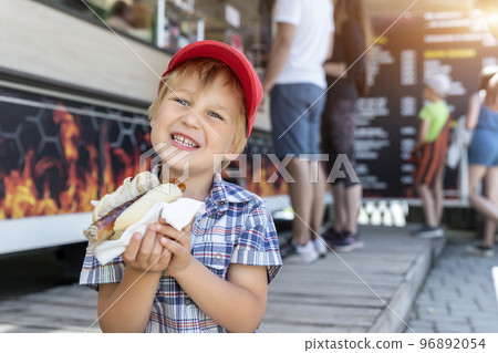 Cute adorable little happy smiling boy kid enjoy eating hot dog sausage in bread near street cafe stall outdoors. Child healthy eating lunch hotdog. Junk food and fast food unhealthy snack concept 96892054