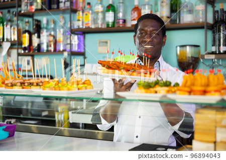 Smiling African American bartender standing with plates of pinchos in front of a pub counter 96894043