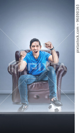 Asian man with the ball sitting on the couch with an excited expression watching football 96898163