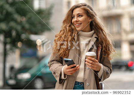 Young woman with curly blonde hair using the phone with a cup of coffee in hands on the city streets Young woman with curly blonde hair using the phone with a cup of coffee in hands on the city streets 96902352
