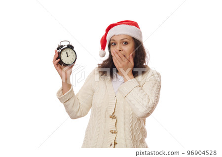Astonished pretty woman in Santa hat, holding a black vintage alarm clock with five minutes to midnight on dial, expecting the upcoming New Year, looking at camera,isolated over white background 96904528