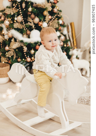 A blonde girl in a sweater rides a wooden horse at home against the background of the Christmas tree. Cute little girl playing at home on the background of garlands. A blonde girl in a sweater rides a wooden horse at home against the background of the Christmas tree. Cute little girl playing at home on the background of garlands. 96907485