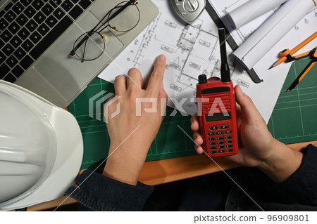 Above view of engineer man hand holding walkie talkie, working on blueprint at wooden office desk 96909801