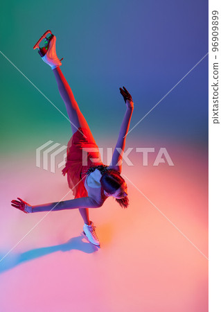 Aerial view of sportive teen girl, junior female figure skater in red stage costume skating isolated over gradient green-blue background in neon light. Grace, beauty, winter sports 96909899